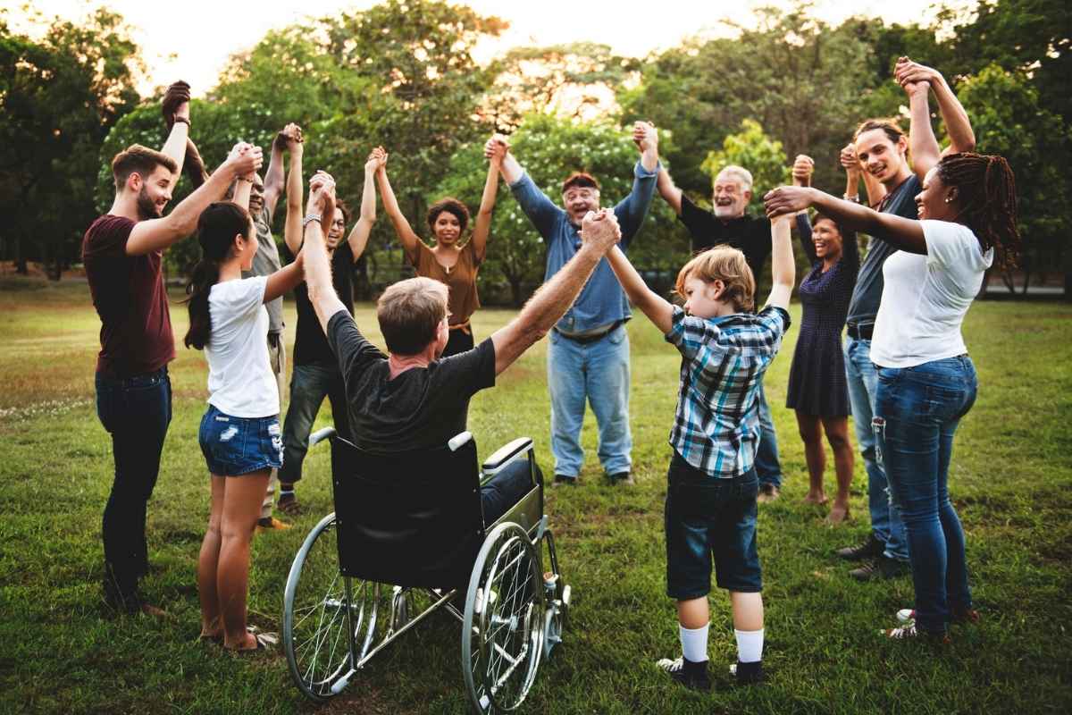 Eine Gruppe von Menschen unterschiedlichen Alters steht im Kreis auf einer Wiese, hält sich an den Händen und hebt gemeinsam die Arme. In der Mitte sitzt eine Person im Rollstuhl.
