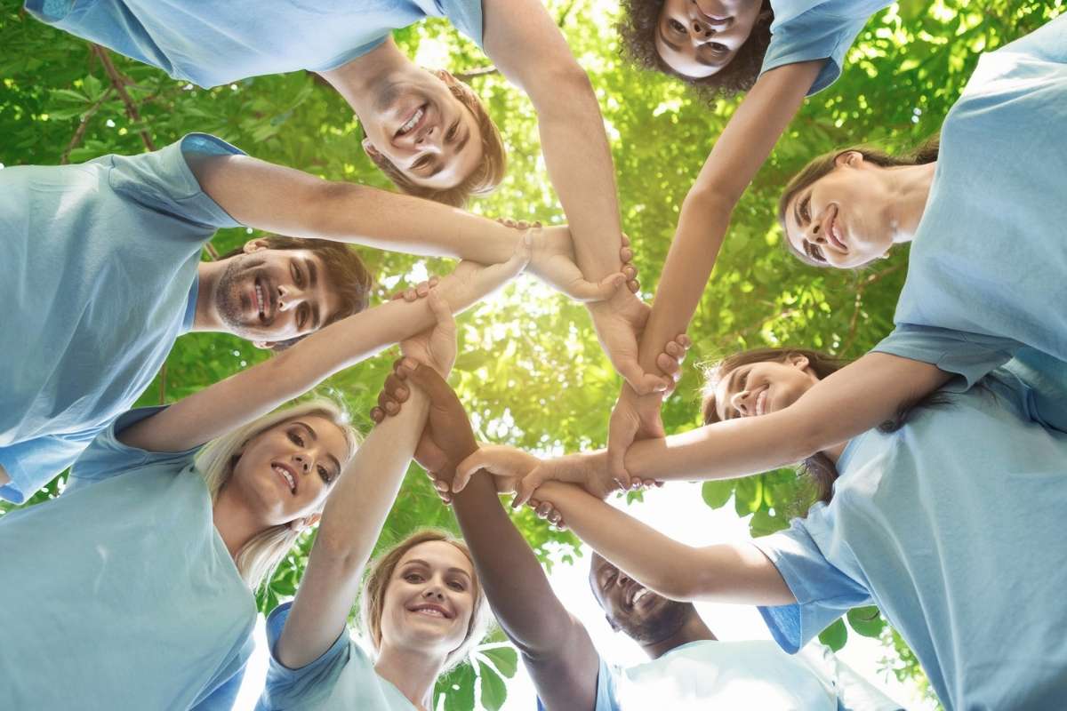 Eine Gruppe von Menschen steht im Kreis unter grünen Bäumen und legt die Hände in der Mitte übereinander als Zeichen für Gemeinschaft und Zusammenhalt.