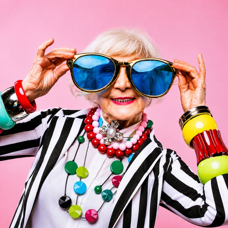 **ALT-Text:** Ältere Frau mit weißen Haaren hält eine übergroße Sonnenbrille mit blauen Gläsern vor ihr Gesicht. Sie trägt eine schwarz-weiß gestreifte Jacke, bunte Schmuckketten und auffällige Armreifen. Der rosa Hintergrund und die farbenfrohen Accessoires vermitteln einen fröhlichen, modernen und selbstbewussten Eindruck.